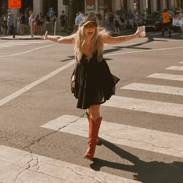 A woman wearing a black dress and Justin Western boots walking down the street while spreading her arms.